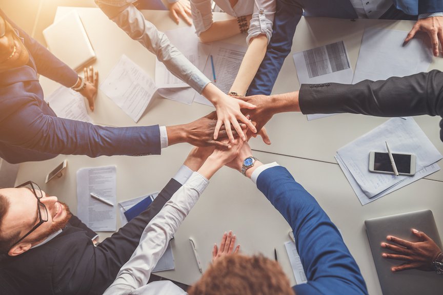 Top-down view of a diverse group of business professionals standing around a conference table, stacking their hands together in the center over documents, laptops, and a smartphone, symbolizing teamwork and collaboration.